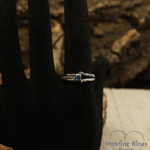 May include: A silver ring with a blue gemstone and a delicate, twisted band. The ring is set on a black velvet background.