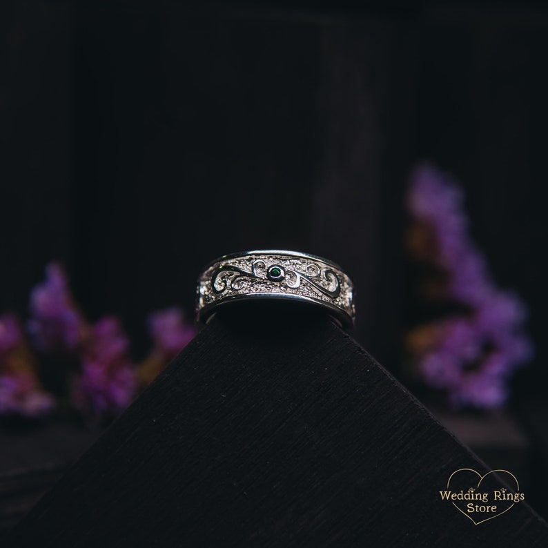 May include: A silver wedding band with an intricate design featuring a green gemstone. The band is on a black surface.