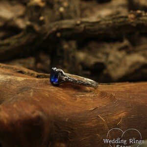May include: A silver ring with a blue oval-shaped gemstone set in a detailed, intricate design. The ring is resting on a piece of wood.