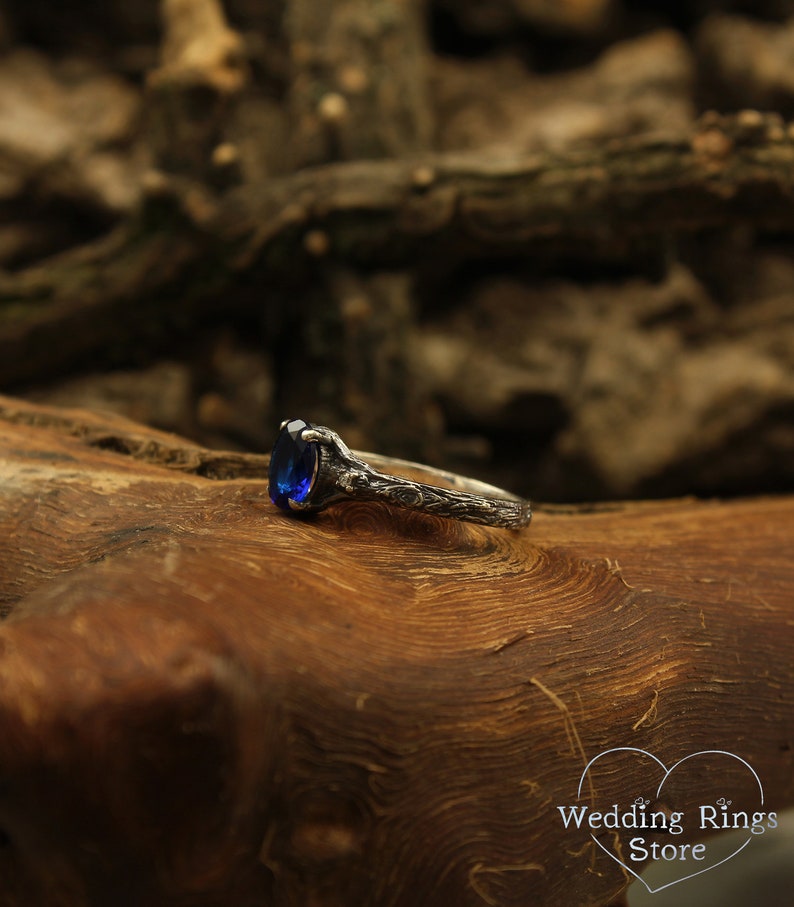 May include: A silver ring with a blue oval-shaped gemstone set in a detailed, intricate design. The ring is resting on a piece of wood.