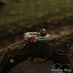 May include: A silver engagement ring with a large red gemstone surrounded by smaller white gemstones. The ring is set on a branch.