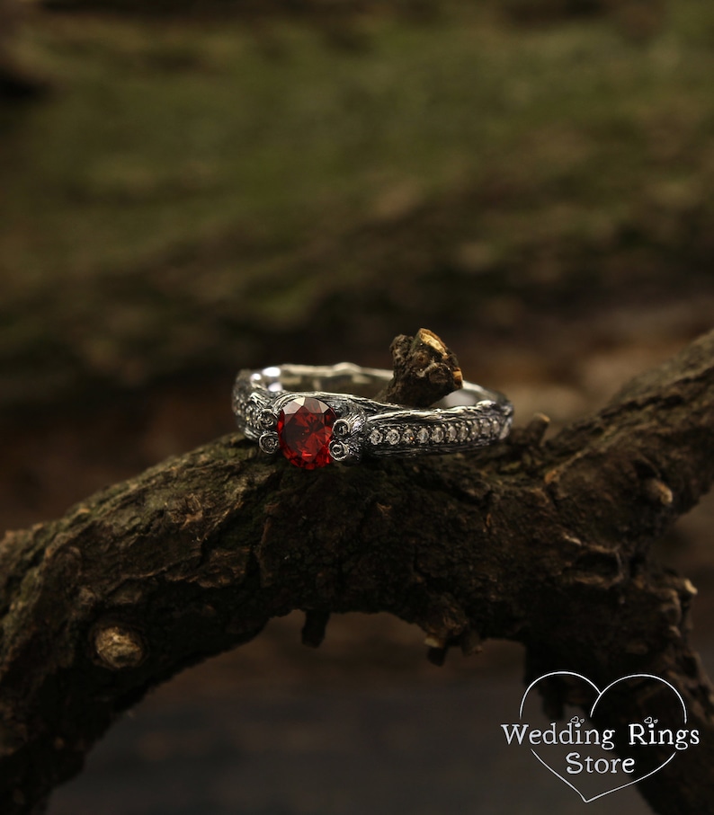 May include: A silver engagement ring with a large red gemstone surrounded by smaller white gemstones. The ring is set on a branch.