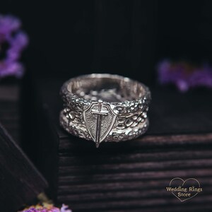 May include: A silver wedding band with a textured design and a shield with a sword on top. The ring is on a dark wooden surface.