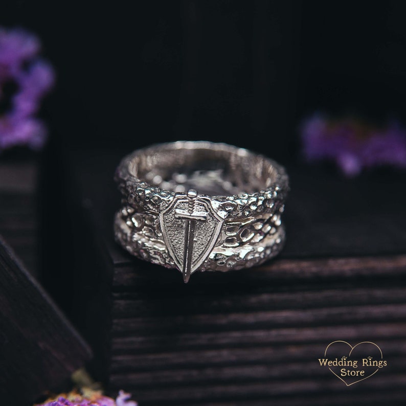 May include: A silver wedding band with a textured design and a shield with a sword on top. The ring is on a dark wooden surface.