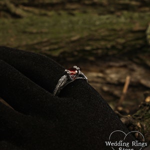 May include: A silver ring with a red gemstone and intricate details, resembling branches or vines. The ring is on a black velvet surface.