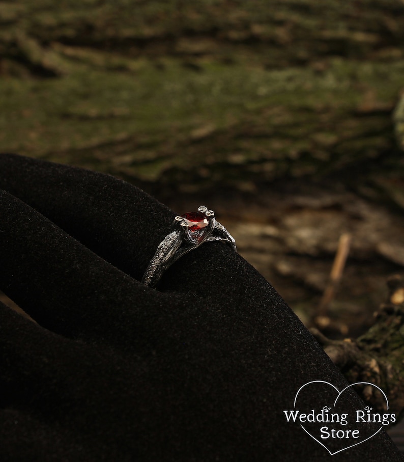 May include: A silver ring with a red gemstone and intricate details, resembling branches or vines. The ring is on a black velvet surface.