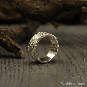 May include: A silver wedding band with an intricate, ornate design. The band is smooth and polished, with a delicate pattern of swirls and curves.
