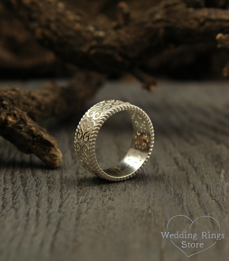 May include: A silver wedding band with an intricate, ornate design. The band is smooth and polished, with a delicate pattern of swirls and curves.