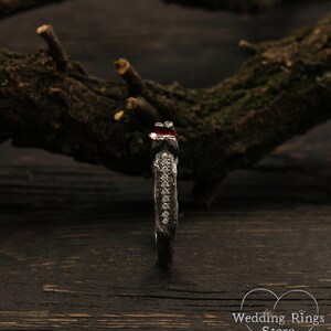 May include: A black oxidized silver ring with a red gemstone and a row of small clear gemstones. The ring is set on a dark wooden surface.