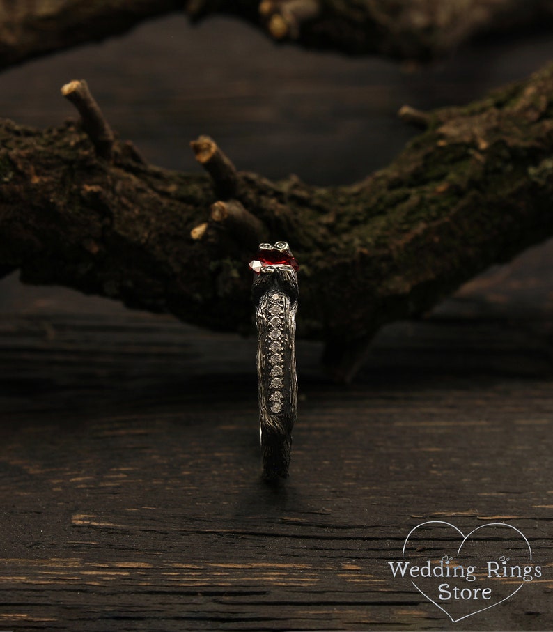 May include: A black oxidized silver ring with a red gemstone and a row of small clear gemstones. The ring is set on a dark wooden surface.