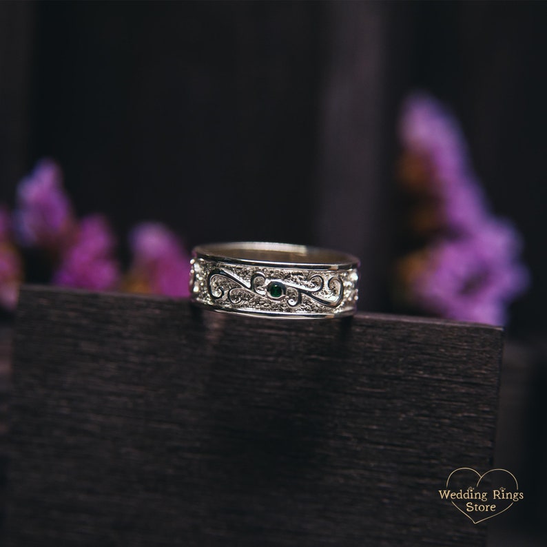 May include: A silver wedding band with a delicate, intricate design and a small green gemstone in the center.
