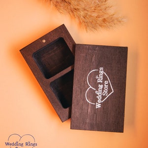 May include: A dark brown wooden ring box with two compartments. The lid has a white heart with the text "Wedding Rings Store" engraved on it.