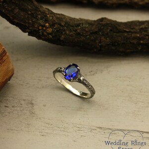 May include: A silver ring with a blue oval gemstone set in a delicate, twig-like band. The ring is on a light wood background.