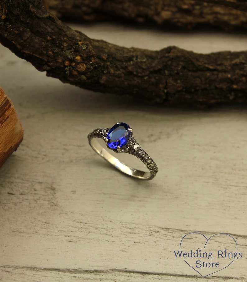May include: A silver ring with a blue oval gemstone set in a delicate, twig-like band. The ring is on a light wood background.