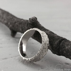 May include: A silver wedding band with a textured bark-like design. The band is resting on a branch.