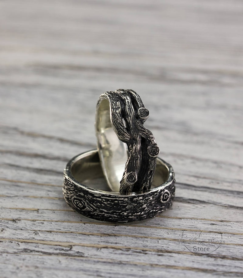May include: Two silver rings with a textured wood-like design. The top ring features a twisted branch design with small black gemstones. The bottom ring has a more subtle wood grain pattern.