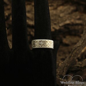 May include: A silver wedding band with a delicate, intricate design featuring a repeating pattern of hearts and swirls. The band is set with small, clear gemstones.