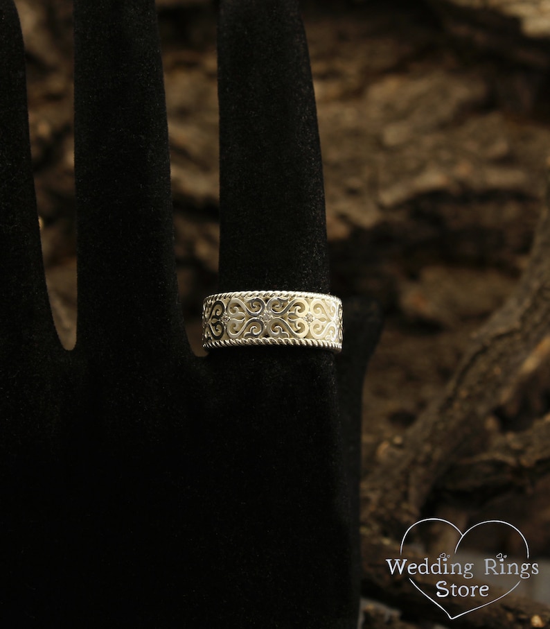 May include: A silver wedding band with a delicate, intricate design featuring a repeating pattern of hearts and swirls. The band is set with small, clear gemstones.