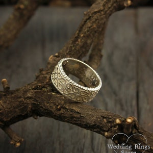 May include: A silver wedding band with a detailed, intricate design of swirling patterns. The band is resting on a branch of a tree.