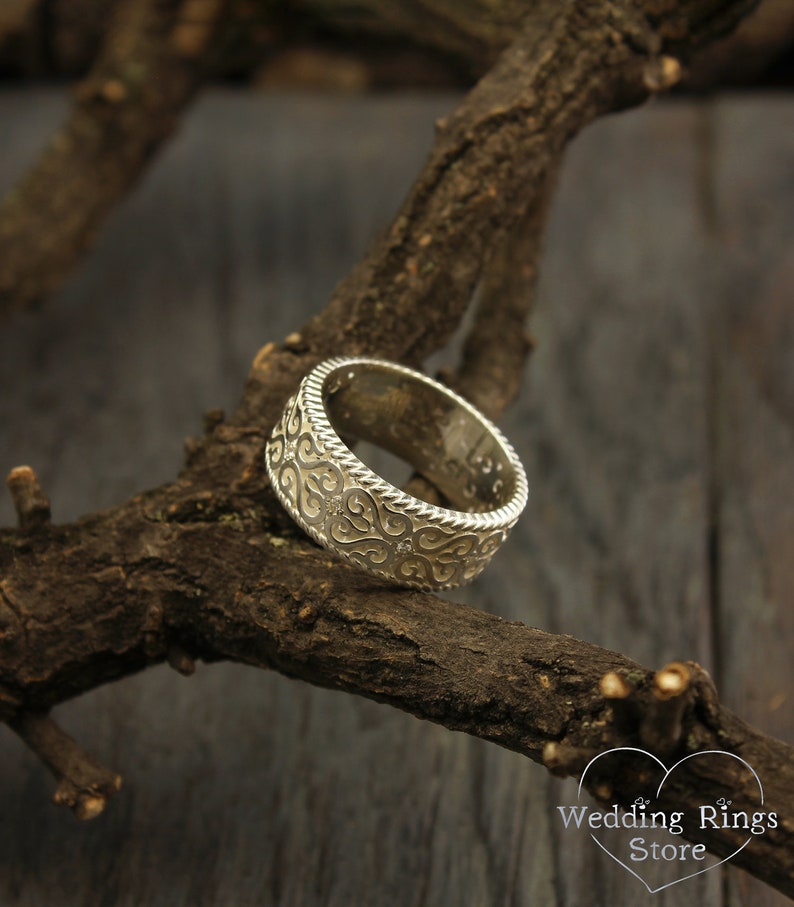 May include: A silver wedding band with a detailed, intricate design of swirling patterns. The band is resting on a branch of a tree.