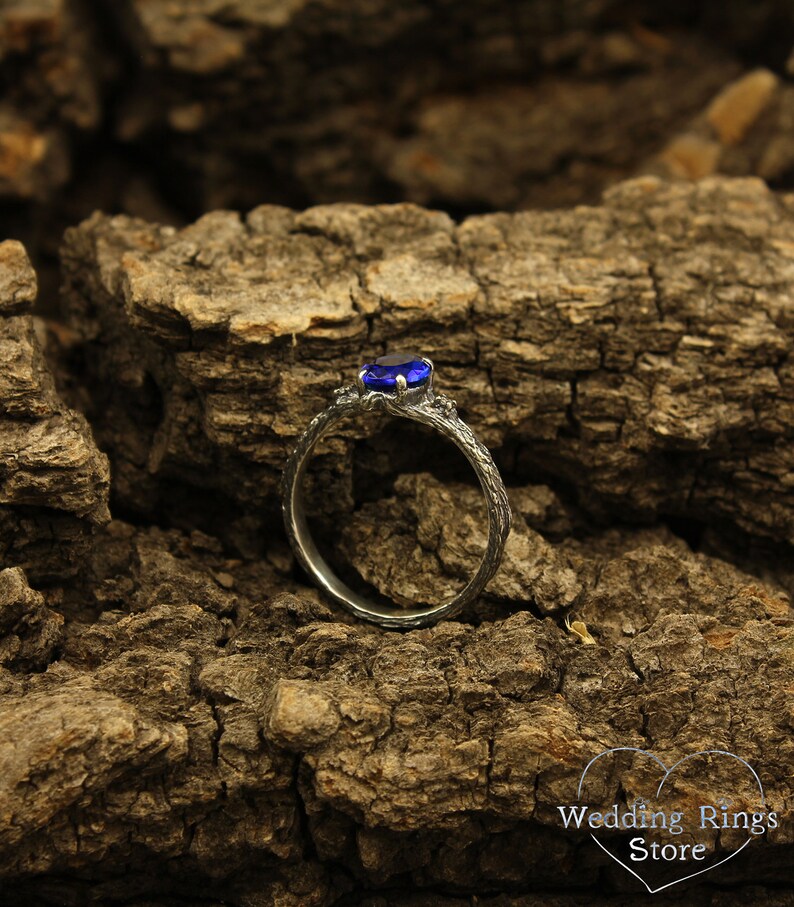 May include: A silver ring with a blue gemstone set in a branch-like design. The ring is on a rough, brown, textured surface.