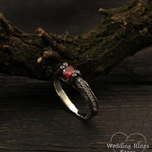 May include: A black and silver ring with a red gemstone and small diamonds set in the band. The ring is on a dark wooden surface.
