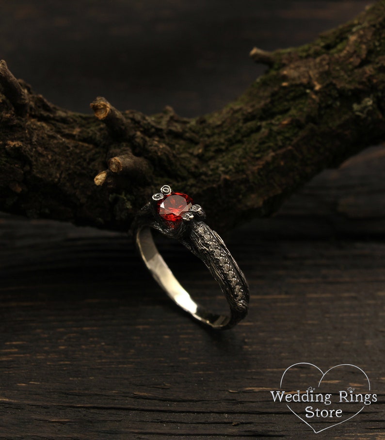 May include: A black and silver ring with a red gemstone and small diamonds set in the band. The ring is on a dark wooden surface.
