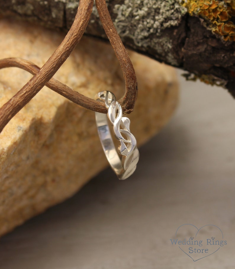 May include: A silver wedding band with a braided design. The band is on a rustic background.