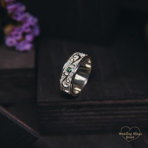 May include: A silver wedding band with an intricate design and a small green gemstone. The band is on a dark wooden surface.
