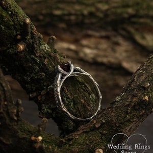 May include: A silver ring with a dark red gemstone set in a branch-like design. The ring is resting on a mossy tree branch.