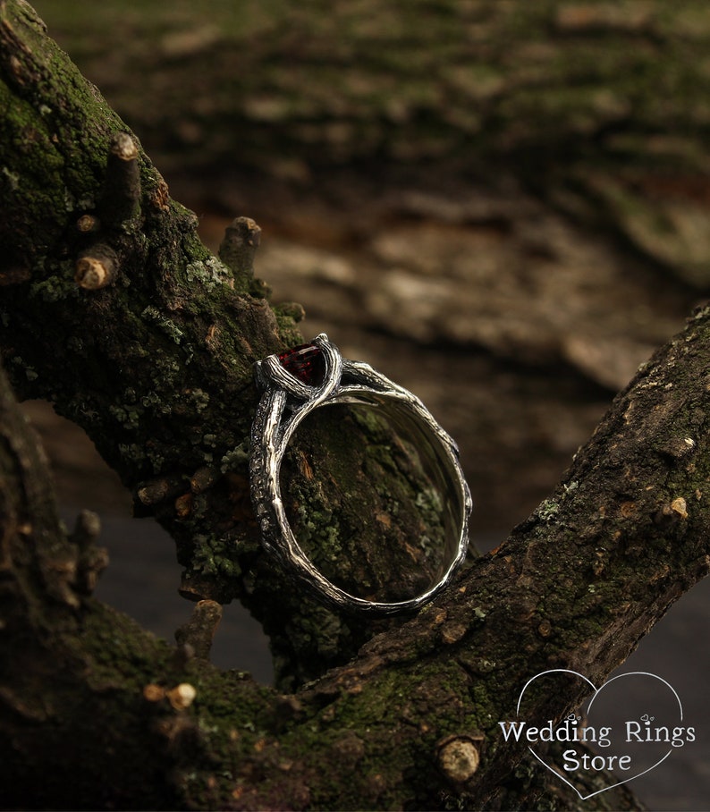 May include: A silver ring with a dark red gemstone set in a branch-like design. The ring is resting on a mossy tree branch.