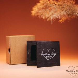 May include: A wooden ring box with a heart-shaped design and the text "Wedding Rings Store" on the lid. The box is closed and sitting on a brown surface.