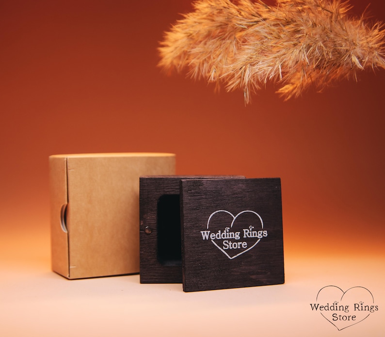 May include: A wooden ring box with a heart-shaped design and the text "Wedding Rings Store" on the lid. The box is closed and sitting on a brown surface.