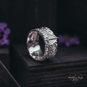 May include: A silver wedding band with a textured design and a small shield-shaped emblem. The band is on a black surface.