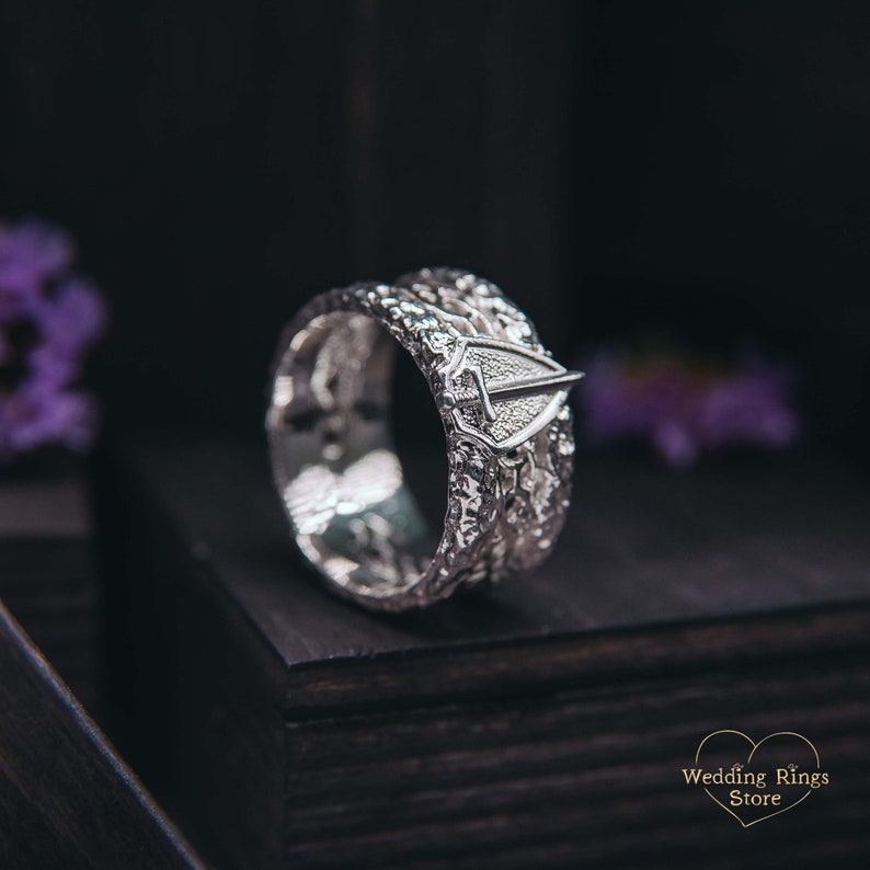 May include: A silver wedding band with a textured design and a small shield-shaped emblem. The band is on a black surface.