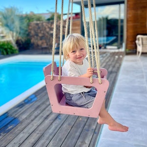 Può includere: Un bambino piccolo è seduto su un'altalena di legno rosa con cinghie di corda. L'altalena è di fronte a una piscina e una casa.