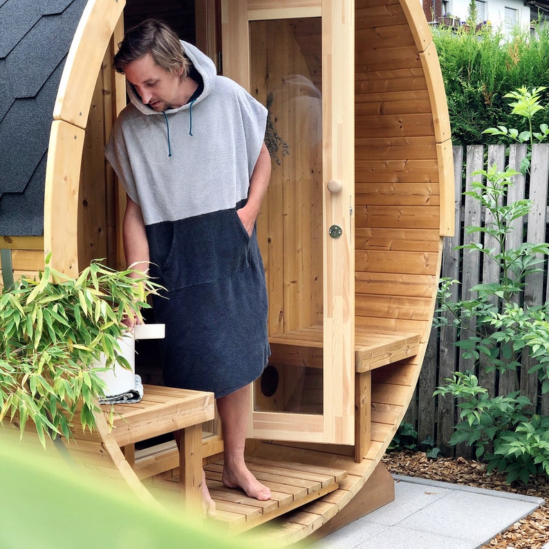 May include: A person wearing a grey and black hooded towel robe stands in the doorway of a wooden barrel sauna. The sauna is made of natural wood and has a wooden door with a glass window. The sauna is surrounded by green plants.