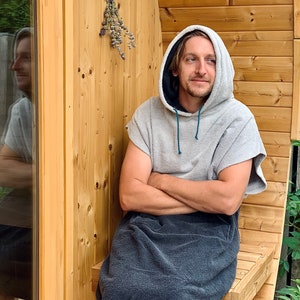 May include: A person wearing a grey hooded towel robe sits on a wooden bench in front of a wooden sauna. The person has their arms crossed and is looking to the right.
