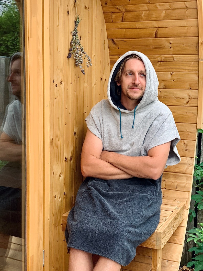 May include: A person wearing a grey hooded towel robe sits on a wooden bench in front of a wooden sauna. The person has their arms crossed and is looking to the right.