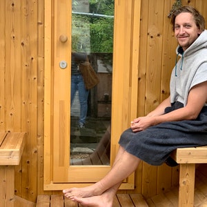 May include: A person wearing a grey hooded towel sits on a wooden bench outside a wooden sauna. The sauna has a glass door and a wooden bench inside.