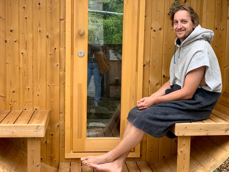 May include: A person wearing a grey hooded towel sits on a wooden bench outside a wooden sauna. The sauna has a glass door and a wooden bench inside.