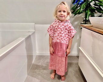 May include: A young person wearing a pink hooded towel with a fish print. The towel has a solid pink bottom half. The person is standing in a bathroom with a white bathtub and white cabinets.