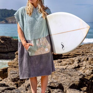K&ouml;nnte beinhalten: Eine Frau tr&auml;gt ein Kapuzenhandtuch in Hellblau und Grau mit einer taschenartigen Karte, h&auml;lt ein wei&szlig;es Surfbrett. Sie steht auf felsigem Gel&auml;nde in der N&auml;he des Ozeans.