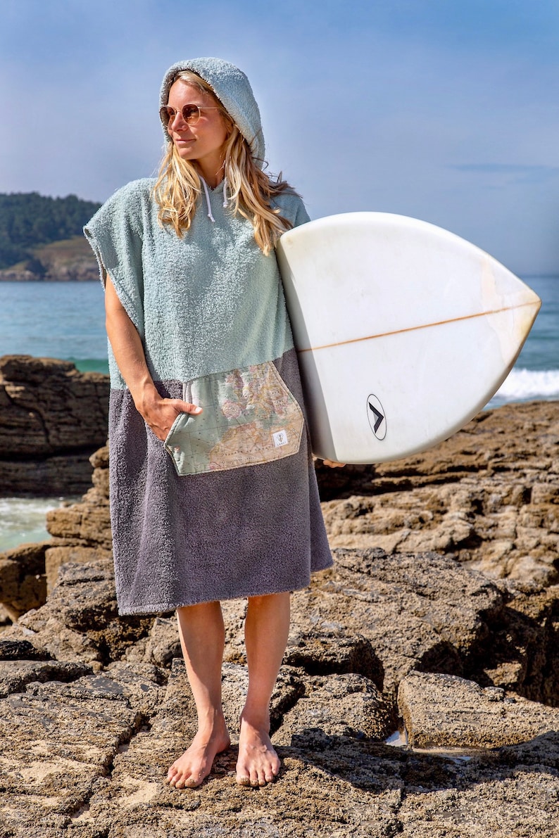 K&ouml;nnte beinhalten: Eine Frau tr&auml;gt ein Kapuzenhandtuch in Hellblau und Grau mit einer taschenartigen Karte, h&auml;lt ein wei&szlig;es Surfbrett. Sie steht auf felsigem Gel&auml;nde in der N&auml;he des Ozeans.