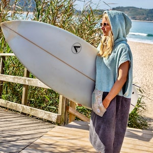 K&ouml;nnte beinhalten: Eine Frau tr&auml;gt ein hellblaues Kapuzenhandtuch mit grauem Unterteil und h&auml;lt ein wei&szlig;es Surfbrett mit einem schwarzen Dreieckslogo. Sie steht auf einem Holzsteg mit einem Strand im Hintergrund.