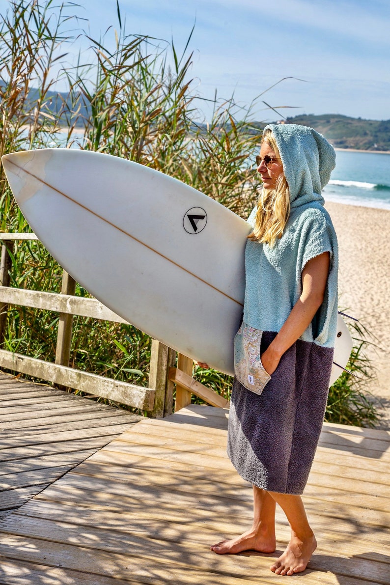 K&ouml;nnte beinhalten: Eine Frau tr&auml;gt ein hellblaues Kapuzenhandtuch mit grauem Unterteil und h&auml;lt ein wei&szlig;es Surfbrett mit einem schwarzen Dreieckslogo. Sie steht auf einem Holzsteg mit einem Strand im Hintergrund.