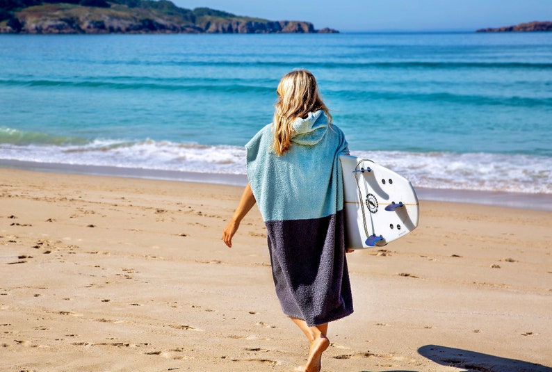 K&ouml;nnte beinhalten: Eine Person geht an einem Sandstrand entlang und tr&auml;gt ein Surfbrett unter dem Arm. Sie tr&auml;gt eine blaue und graue Kapuzenbadetuchrobe.