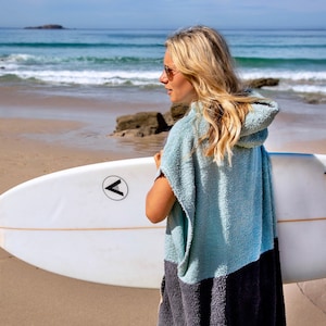 K&ouml;nnte beinhalten: Eine Frau tr&auml;gt ein hellblaues Kapuzenhandtuch mit einem schwarzen Streifen am unteren Rand und h&auml;lt ein wei&szlig;es Surfbrett mit einem schwarzen Pfeil-Logo an der Seite. Das Surfbrett hat blaue Finnen und eine schwarze Leine.