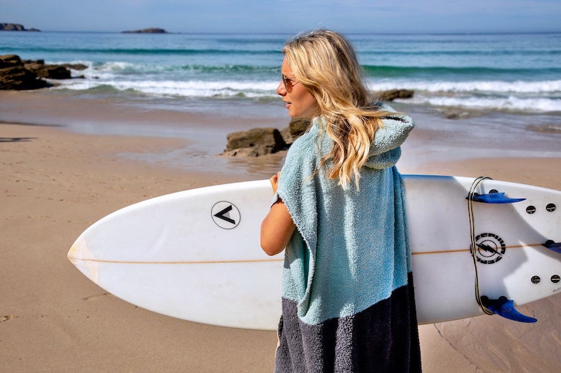 K&ouml;nnte beinhalten: Eine Frau tr&auml;gt ein hellblaues Kapuzenhandtuch mit einem schwarzen Streifen am unteren Rand und h&auml;lt ein wei&szlig;es Surfbrett mit einem schwarzen Pfeil-Logo an der Seite. Das Surfbrett hat blaue Finnen und eine schwarze Leine.