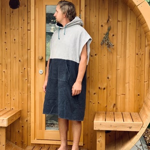May include: A person wearing a grey and black hooded towel robe stands in front of a wooden sauna. The sauna has a wooden door and two benches.